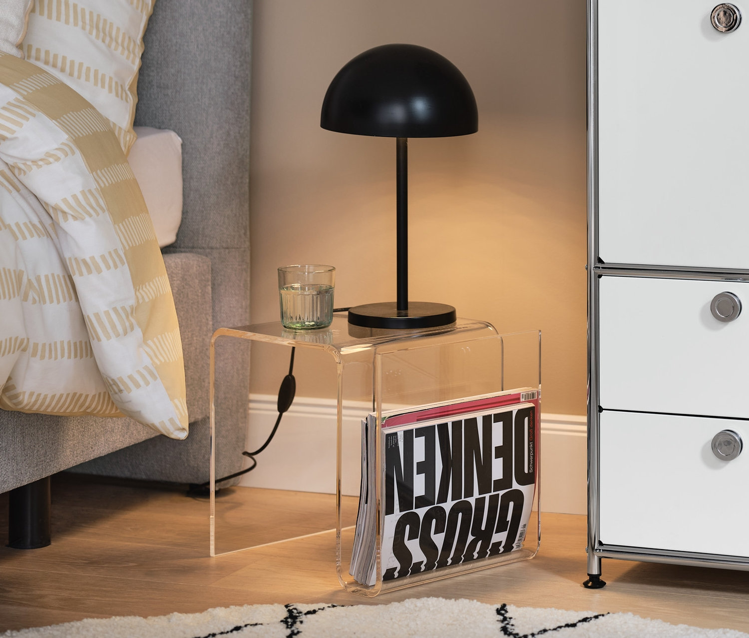Ein Beistelltisch aus Acrylglas mit Magazinhalter steht neben einem Bett. Daneben steht ein Metallschrank CN3 mit Schubladen und Klappen.