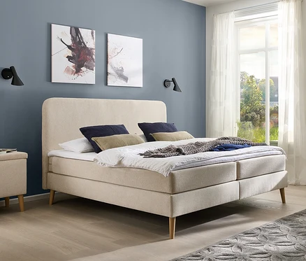 Schlafzimmer mit beigem DOMO collection Boxspringbett mit festem Kopfteil, ca. 180 x 200 cm, inkl. Topper, zwei Nachttischen und grauem Teppich.