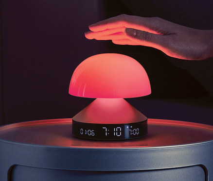 Eine Hand schwebt über einem anthrazitfarbenen LEXON Lichtwecker »Mina Sunrise« auf einem Nachttisch.