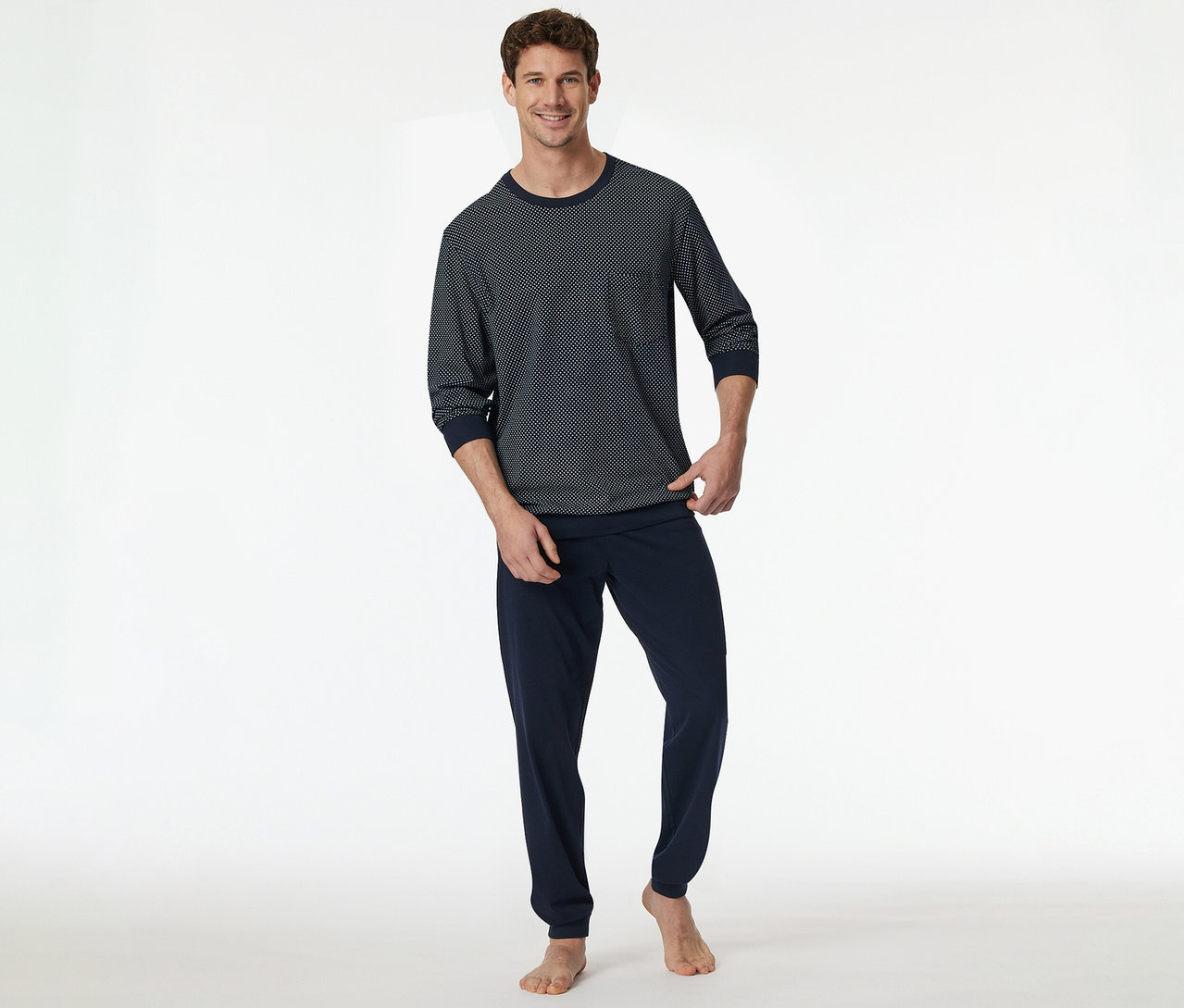 Ein lächelnder Mann steht barfuß und trägt ein SCHIESSER Pyjama-Set. Das Oberteil ist dunkelblau mit weißem geometrischem Muster, die Hose ist dunkelblau.