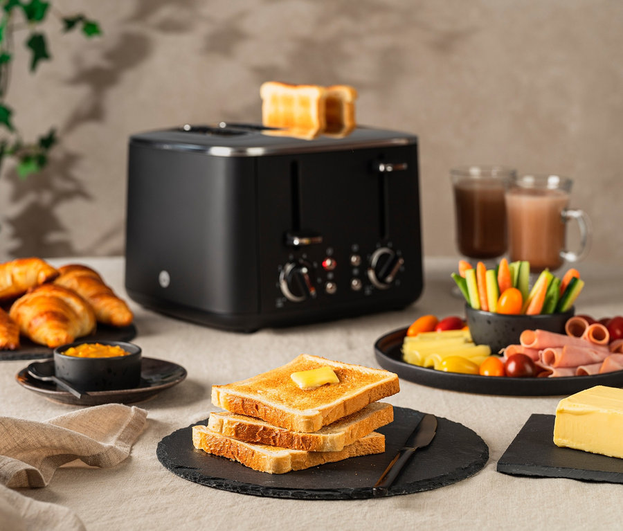 Auf einem Tisch stehen ein schwarzer WILFA Toaster FAMILY, Toast, Croissants, Marmelade, Gemüse, Schinken, Käse, Butter und zwei Getränke.