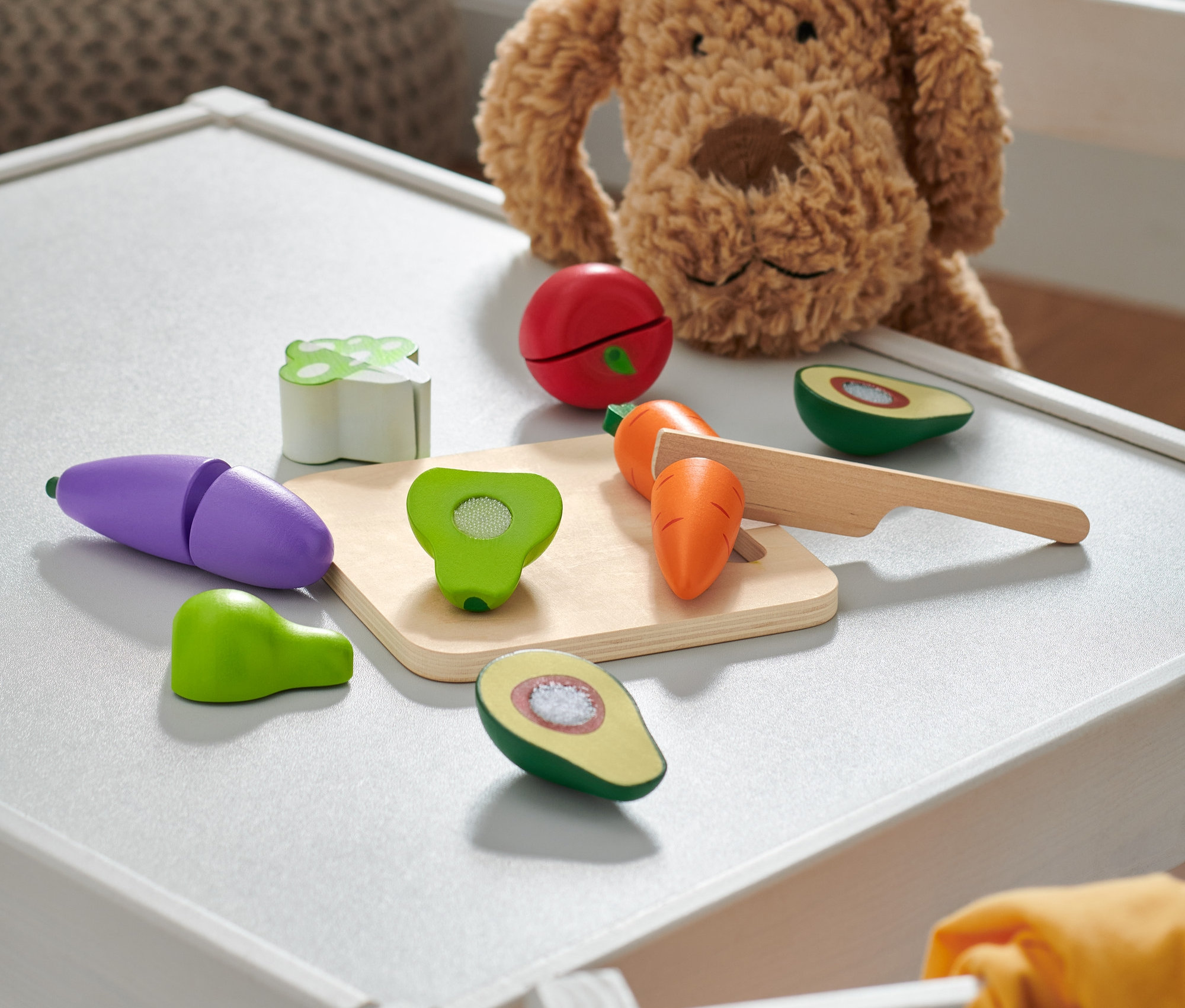 Ein Plüschhund sitzt auf einem weißen Tisch neben hölzernen Spielzeugobst und -gemüse.