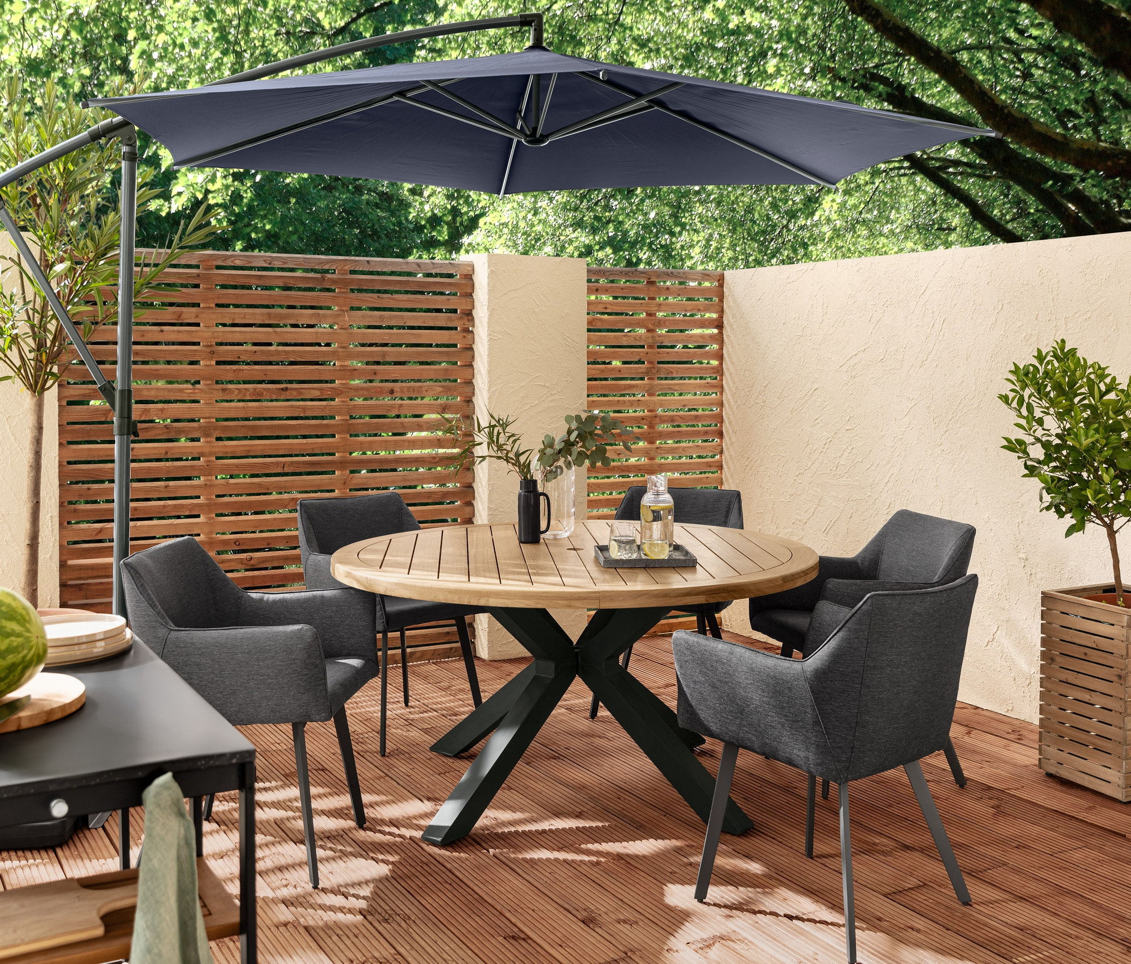 Vier Dining-Sessel mit Quick-Dry-Foam und Outdoor-Tex-Bezug und ein Teak-Dining-Tisch mit modernem X-Fuß stehen auf einer Holzterrasse unter einem Leco Ampelschirm.