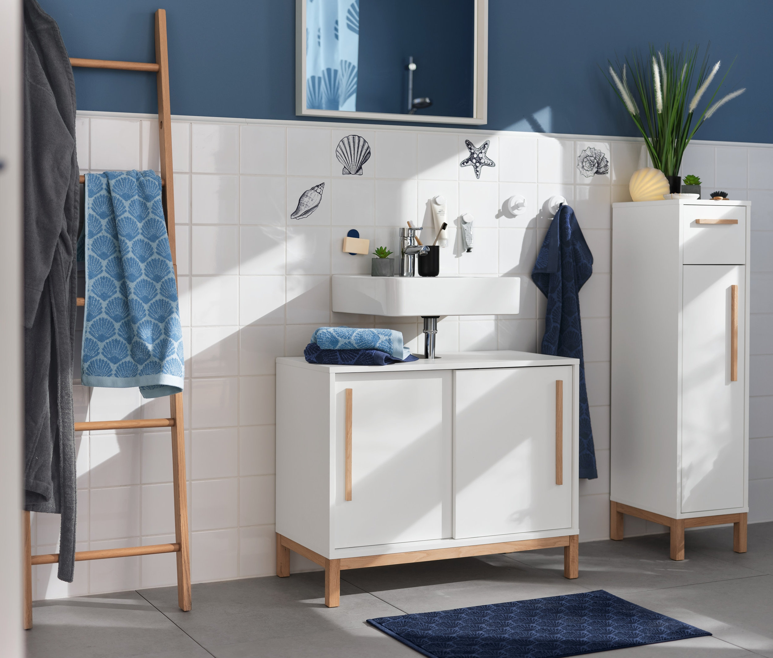 Ein Badezimmer mit Waschbecken, Spiegel und Stauraum. Maritime Fliesenaufkleber schmücken die Wand. Eine Holzleiter hält ein blaues Handtuch »Muschel« und einen grauen Bademantel.
