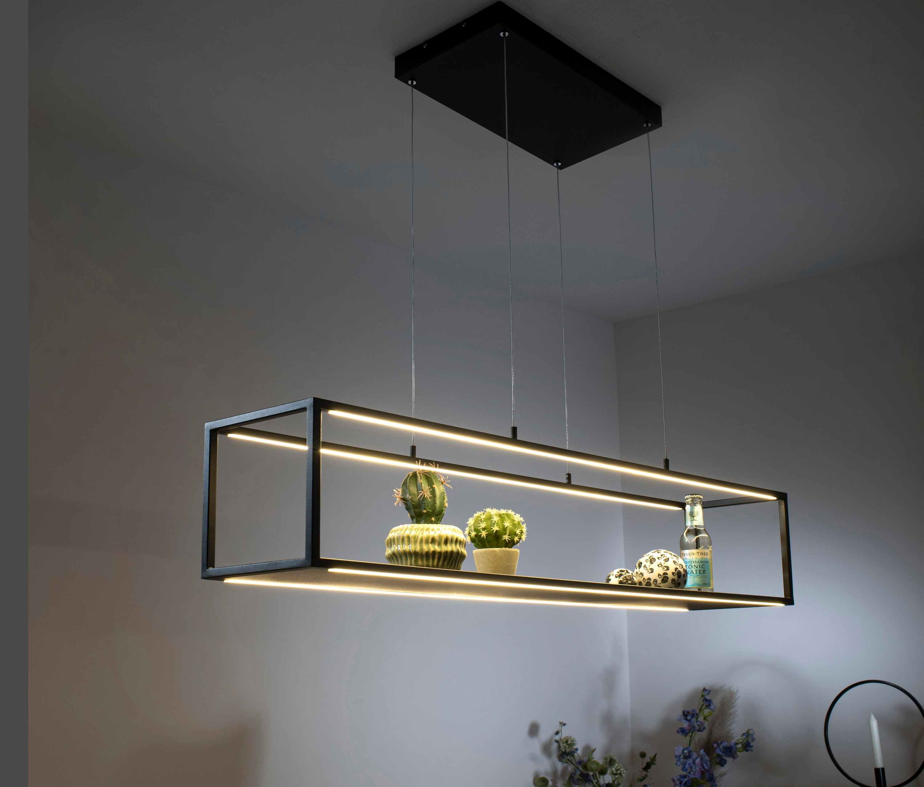 Eine schwarze LED-Pendelleuchte »Contura« hängt über einem Tisch, auf dem Dekorationen und Blumen stehen.