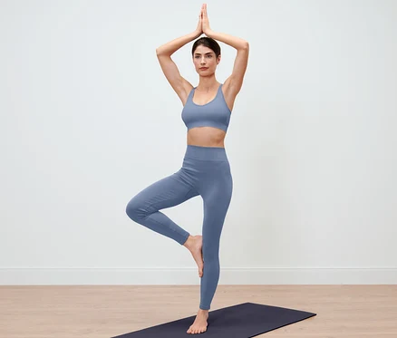 Frau in blauen Seamless-Tight steht in der Baum-Pose auf einer Yogamatte.