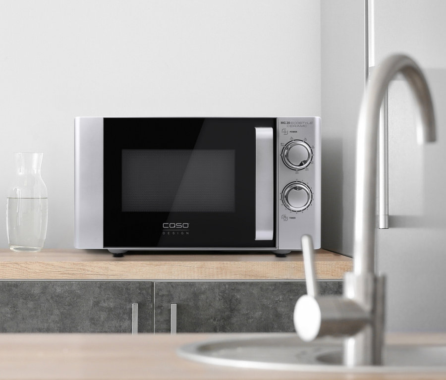 Eine silberne Caso Mikrowelle steht auf einer Küchentheke neben einer Schüssel mit Obst und einer Glaskaraffe mit Wasser.