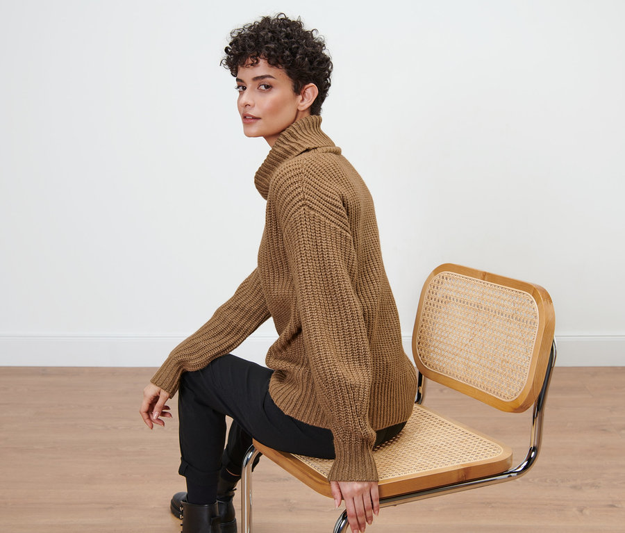 Frau sitzt auf einem Stuhl und trägt einen braunen Grobstrickpullover mit Rollkragen.