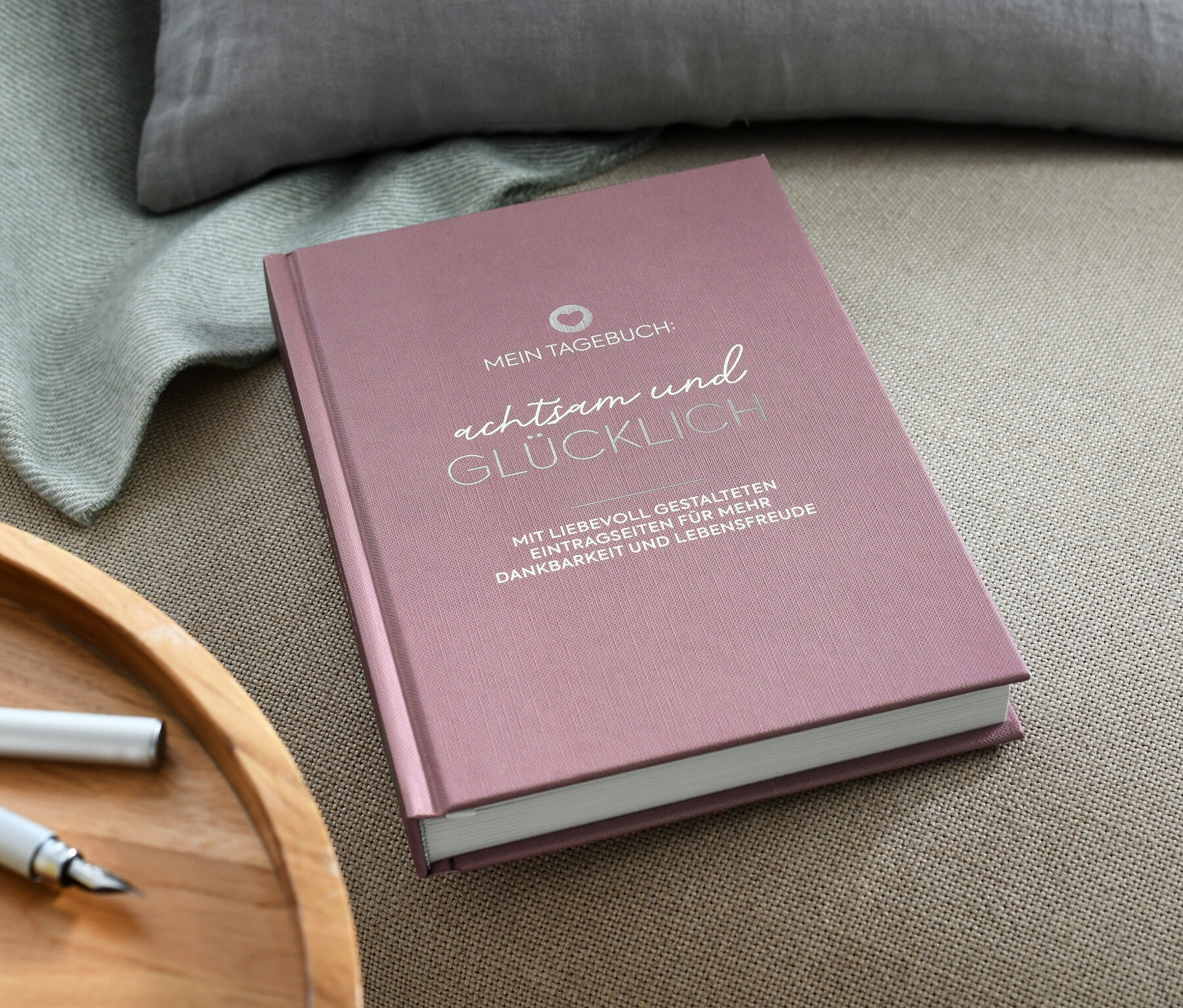 Rosa Buch "Mein Tagebuch - achtsam und Glücklich" liegt auf einem beigen Sessel neben einer Tasse und einem Stift.
