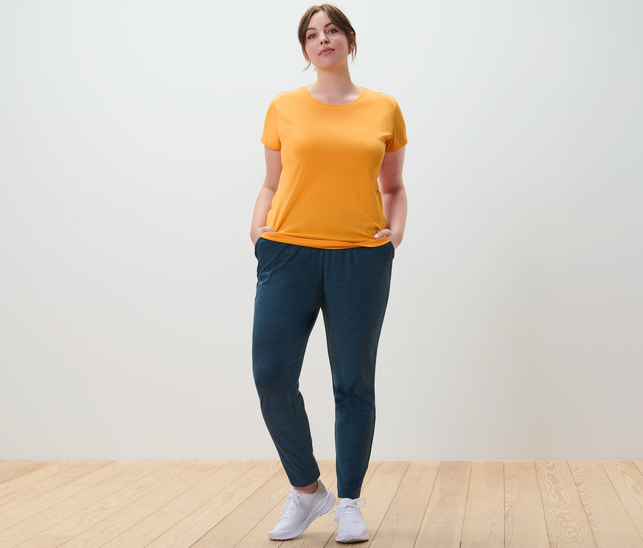 Eine Frau steht mit den Händen in den Taschen ihrer Trainingshose und trägt ein oranges Funktionsshirt.