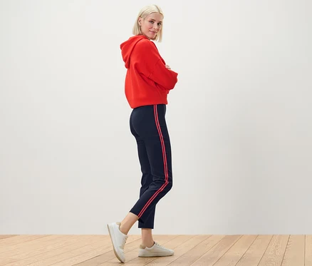 Blonde Frau posiert in rotem Hoodie und Stretchhose mit Galonstreifen.