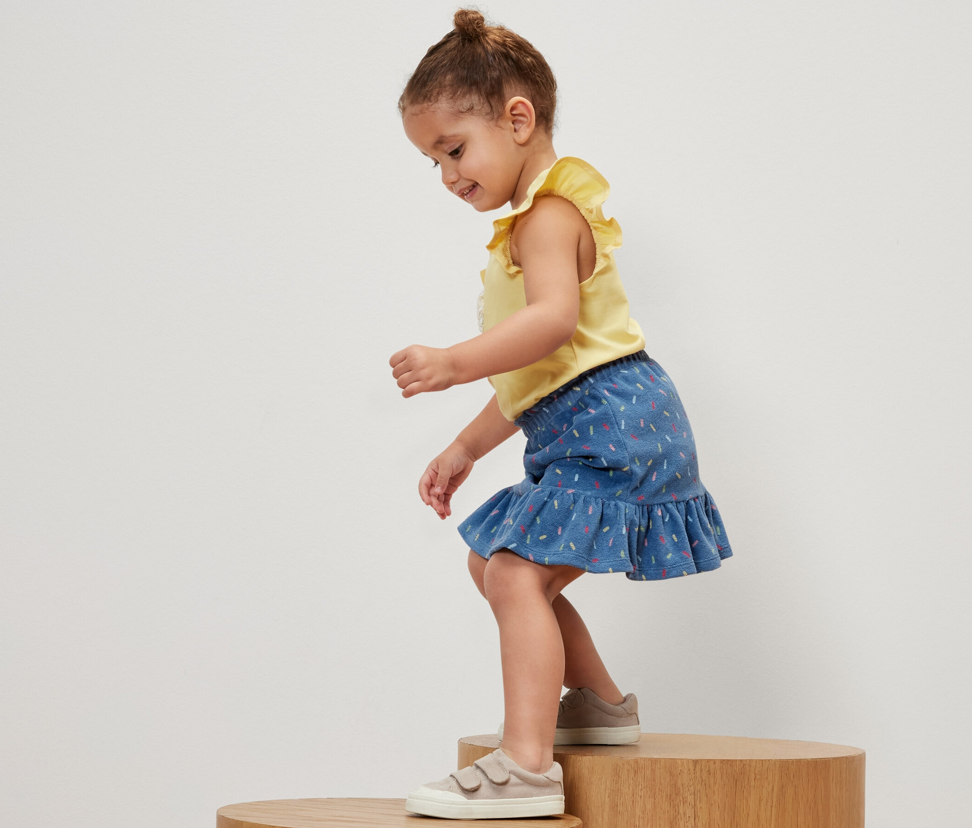 Mädchen steht auf einem Holzsockel und trägt ein gelbes T-Shirt und einen Kinder-Frottier-Rock.
