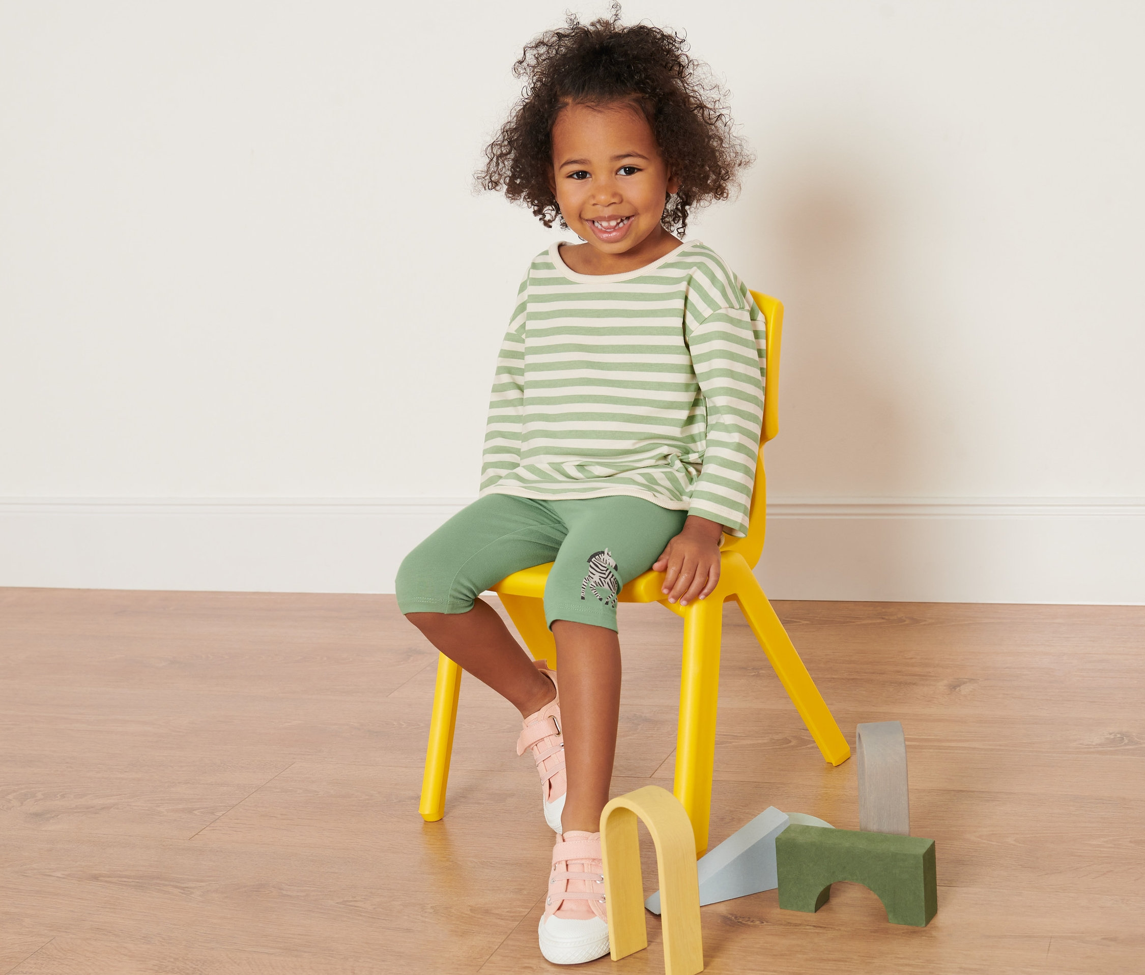 Ein Mädchen sitzt auf einem gelben Stuhl und trägt grüne Leggings mit einem gestreiften Oberteil. Sie trägt 3 Kinder-Leggings.