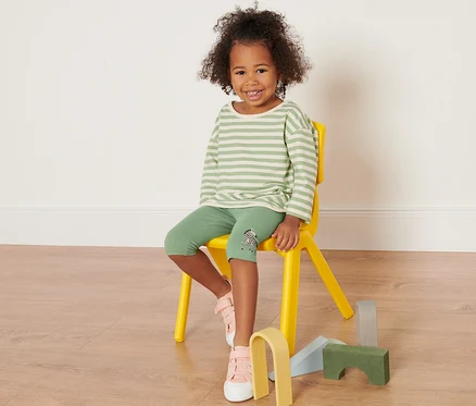 Ein Mädchen sitzt auf einem gelben Stuhl und trägt grüne Leggings mit einem gestreiften Oberteil. Sie trägt 3 Kinder-Leggings.