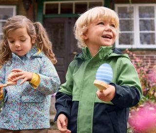 Kinder spielen das Eierlauf-Spiel. Das Mädchen trägt eine Regenjacke mit Blumen und der Junge eine Softshelljacke.