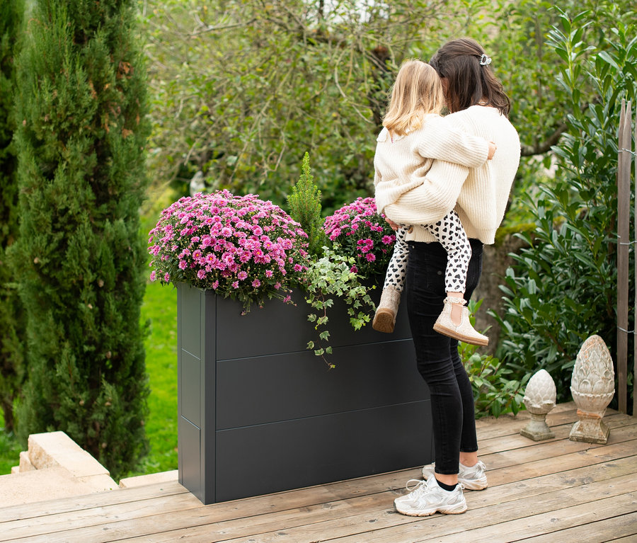 Eine Mutter hält ihre Tochter neben einem grauen Pflanzkasten mit rosa Blumen.