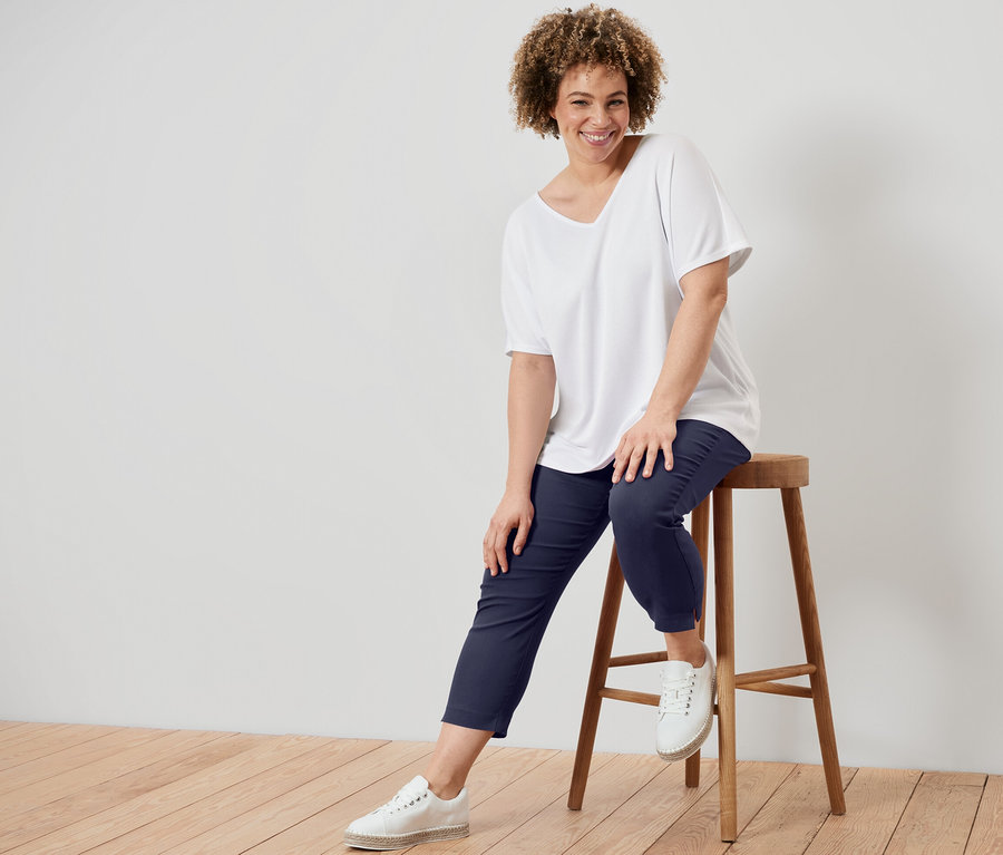 Frau sitzt auf einem Hocker und trägt ein weißes, strukturiertes Shirt und eine dunkelblaue 7/8-Stretchhose.