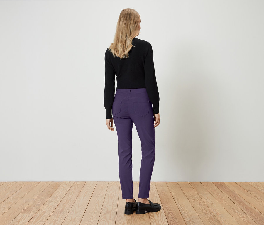 Frau steht mit dem Rücken zur Kamera und trägt ein schwarzes Oberteil und eine violette Hose.