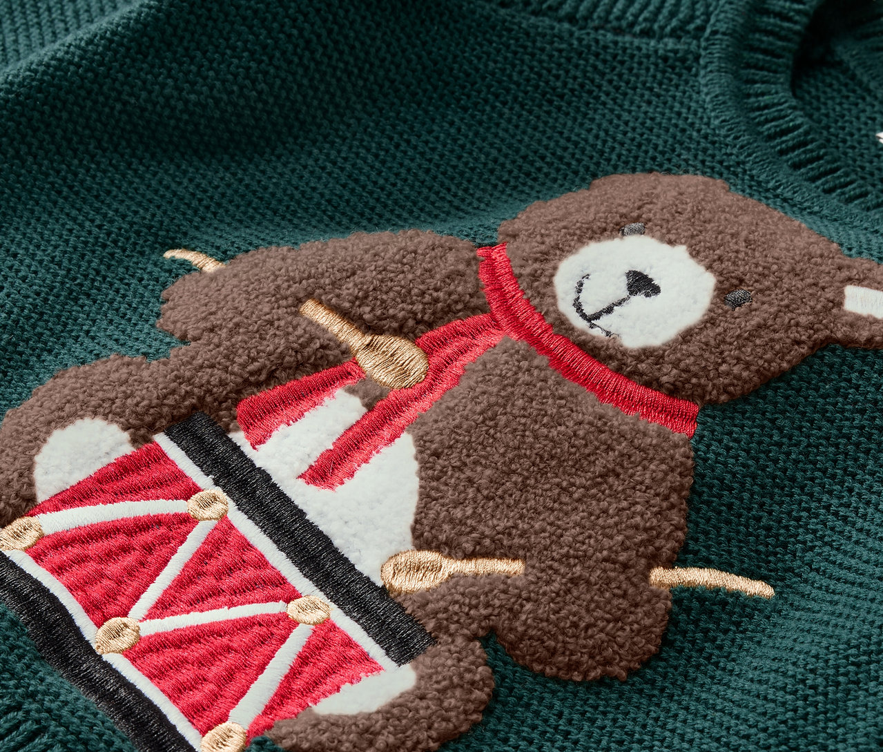Detailaufnahme eines grünen Pullovers mit einer Teddybär-Applikation, der eine Trommel spielt.
