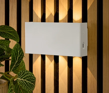 Eine rechteckige, weiße Wandleuchte ist an einer Holzwand befestigt. Darunter liegen Schlüssel auf einem Tisch und eine Blume steht in einem Topf.