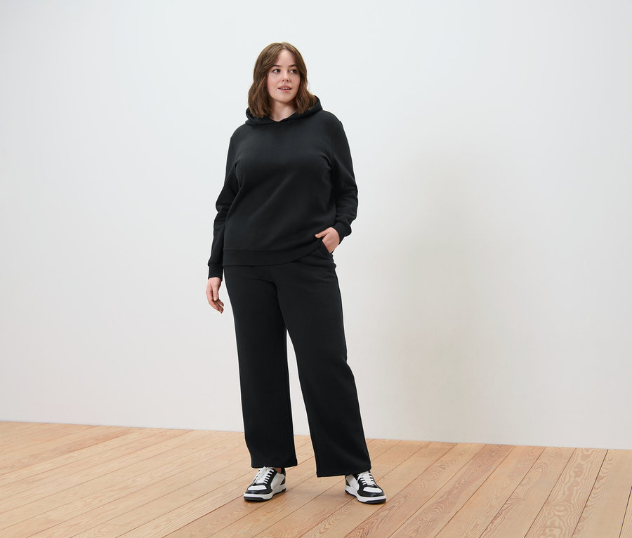 Frau im schwarzen Hoodie-Set mit weiter Hose, stehend auf einem Holzboden.