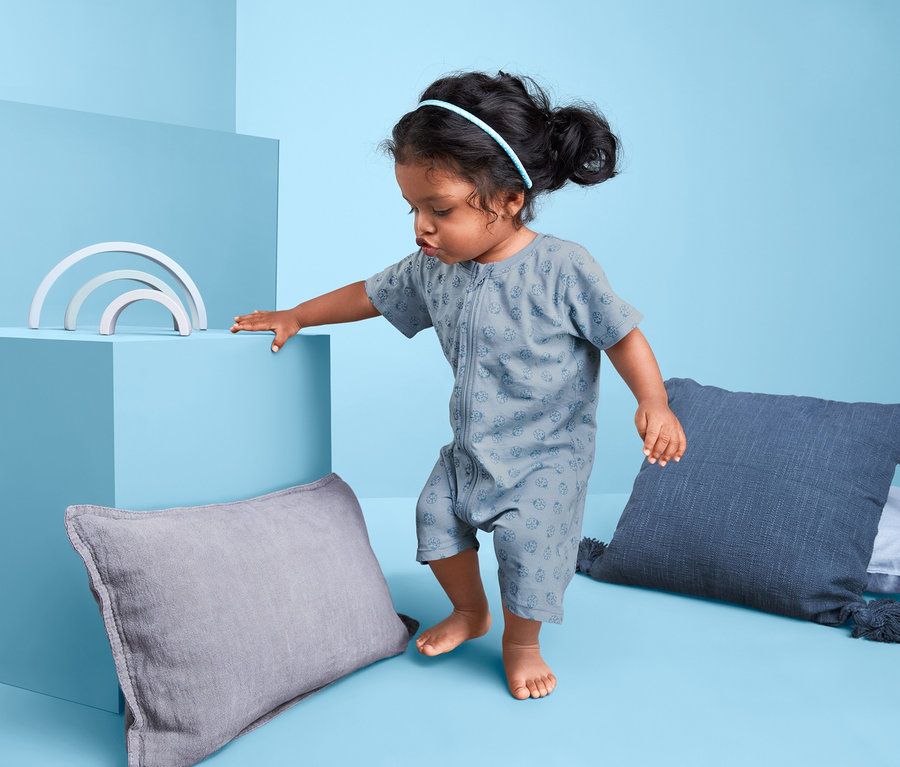 Kleinkind in grün-blauem 2er-Shorty-Pyjama steht neben Kissen und Spielzeug.