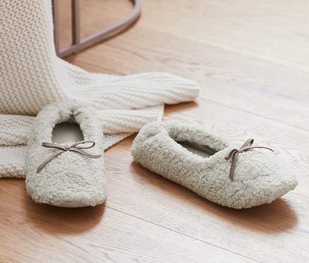 Auf einem Holzboden liegen ein Paar Hausschuhslipper in Creme neben einer weißen Strickdecke und einem rosa Hocker.