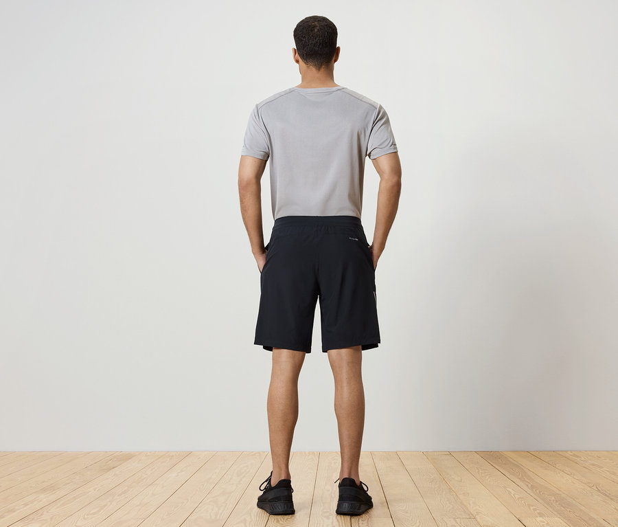 Ein Mann mit dem Rücken zur Kamera trägt ein graues T-Shirt, schwarze Shorts und schwarze Schuhe.