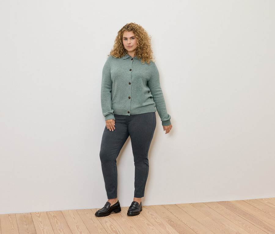 Frau mit lockigem Haar steht und trägt einen grünen Cardigan, graue Hosen und schwarze Slipper.