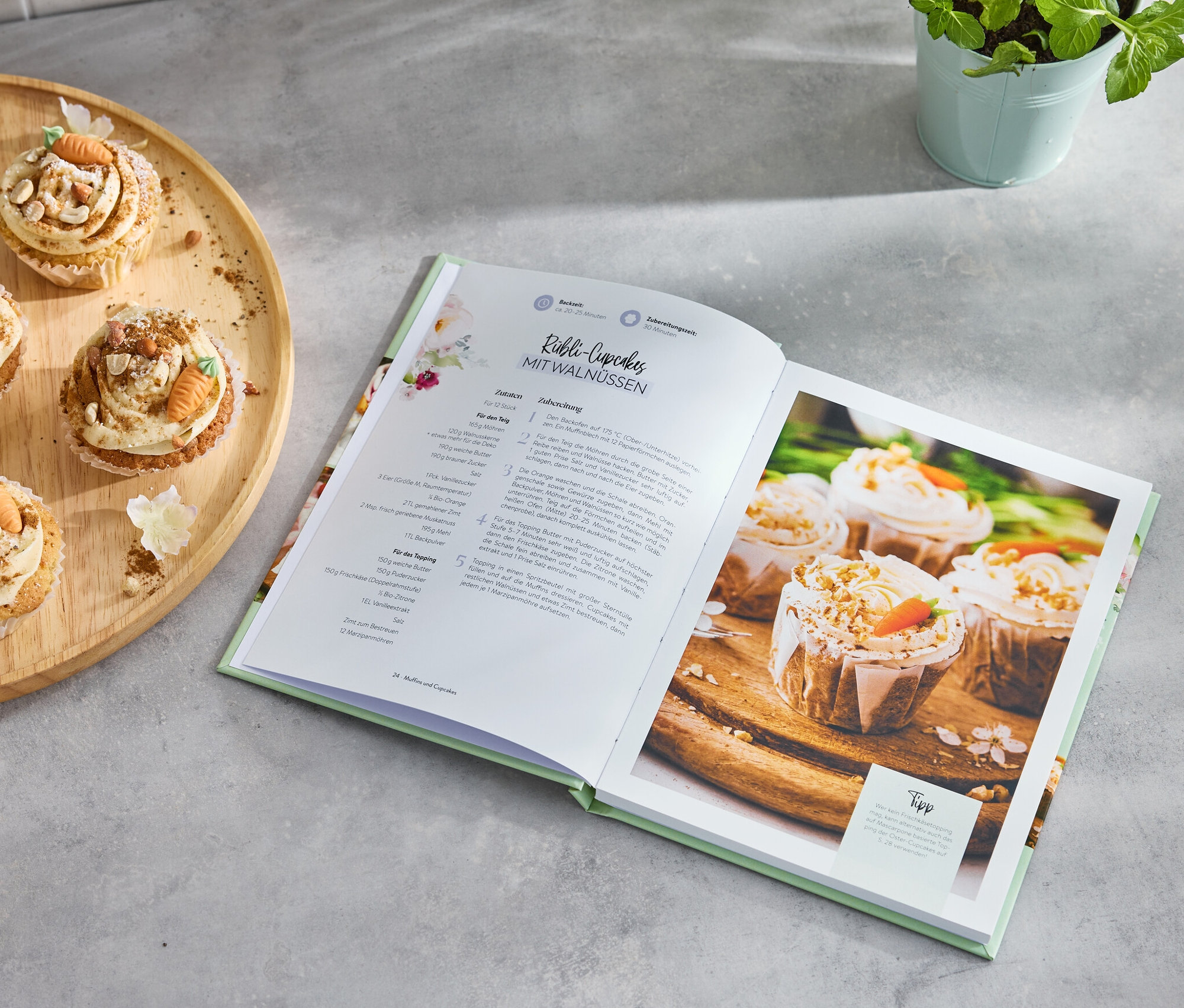 Vier Karotten-Cupcakes auf einem Holztablett neben einem aufgeschlagenen Buch »Kleines Kuchenglück«.