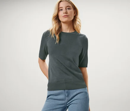 Frau posiert in einem grünen Feinstrickpullover und blauen Jeans.