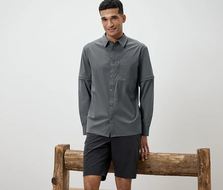 Mann, der sich an eine Holzbank lehnt, trägt ein graues Outdoorhemd und schwarze Shorts.
