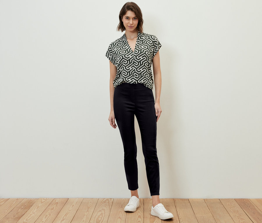 Frau steht und trägt eine schwarze Stretchhose und eine gemusterte Bluse.