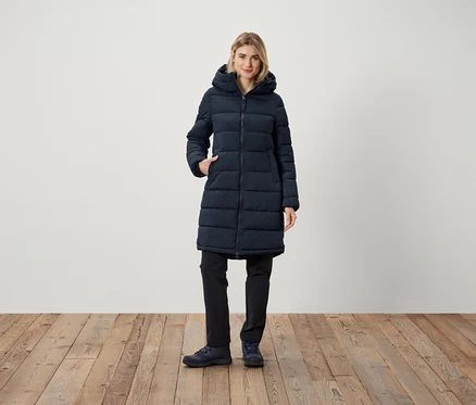 Frau steht mit der Hand in der Tasche einer dunkelblauen Steppjacke mit Kapuze.