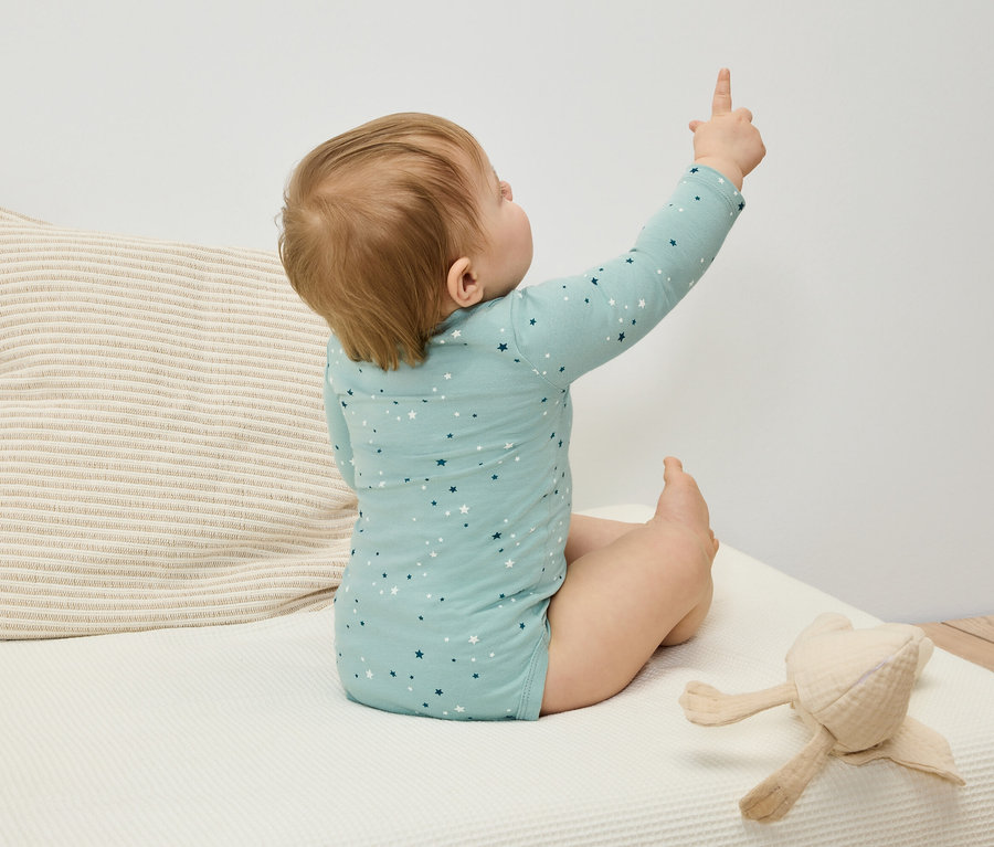 Baby in blauem Sternenbody sitzt auf dem Bett und zeigt mit dem Finger.
