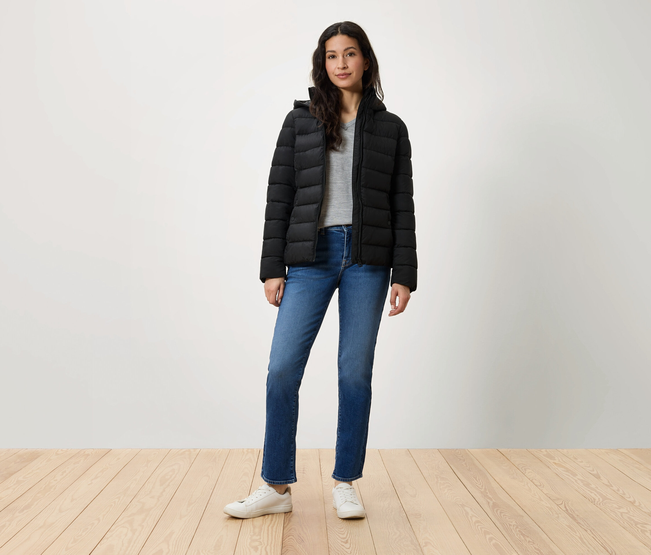 Eine Frau steht und trägt eine schwarze Daunenjacke, blaue Jeans und weiße Turnschuhe.