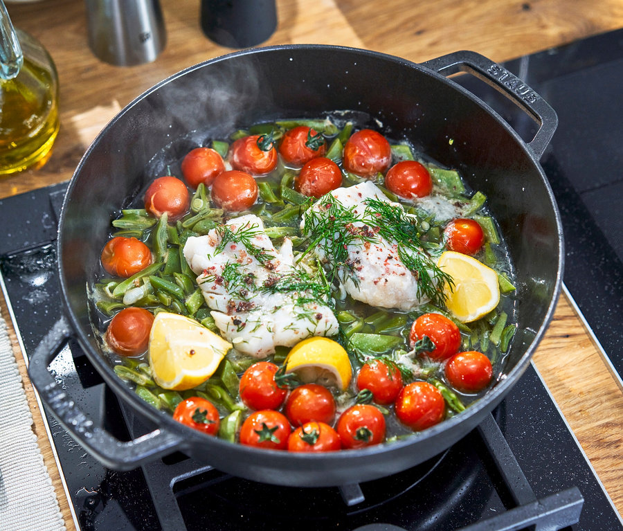 Zwei Fischstücke mit Dill in einem schwarzen Topf mit Kirschtomaten, grünen Bohnen und Zitrone.
