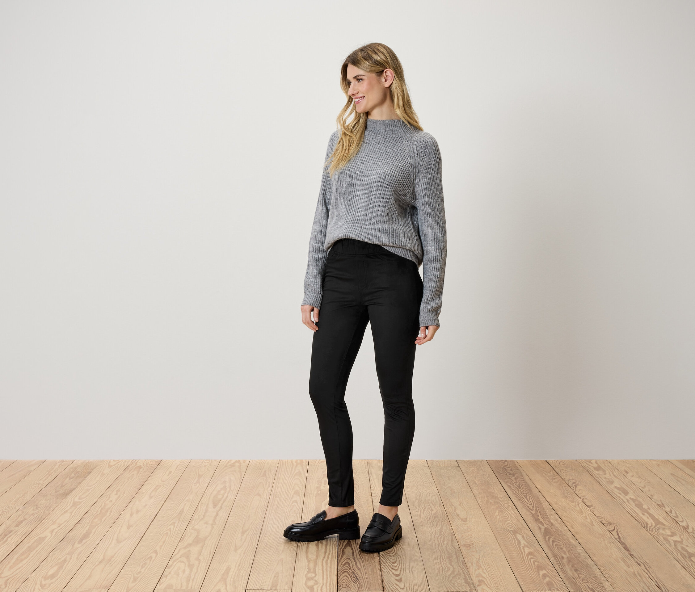 Eine Frau steht auf einem Holzboden und trägt einen grauen Pullover, schwarze Hosen und schwarze Schuhe.