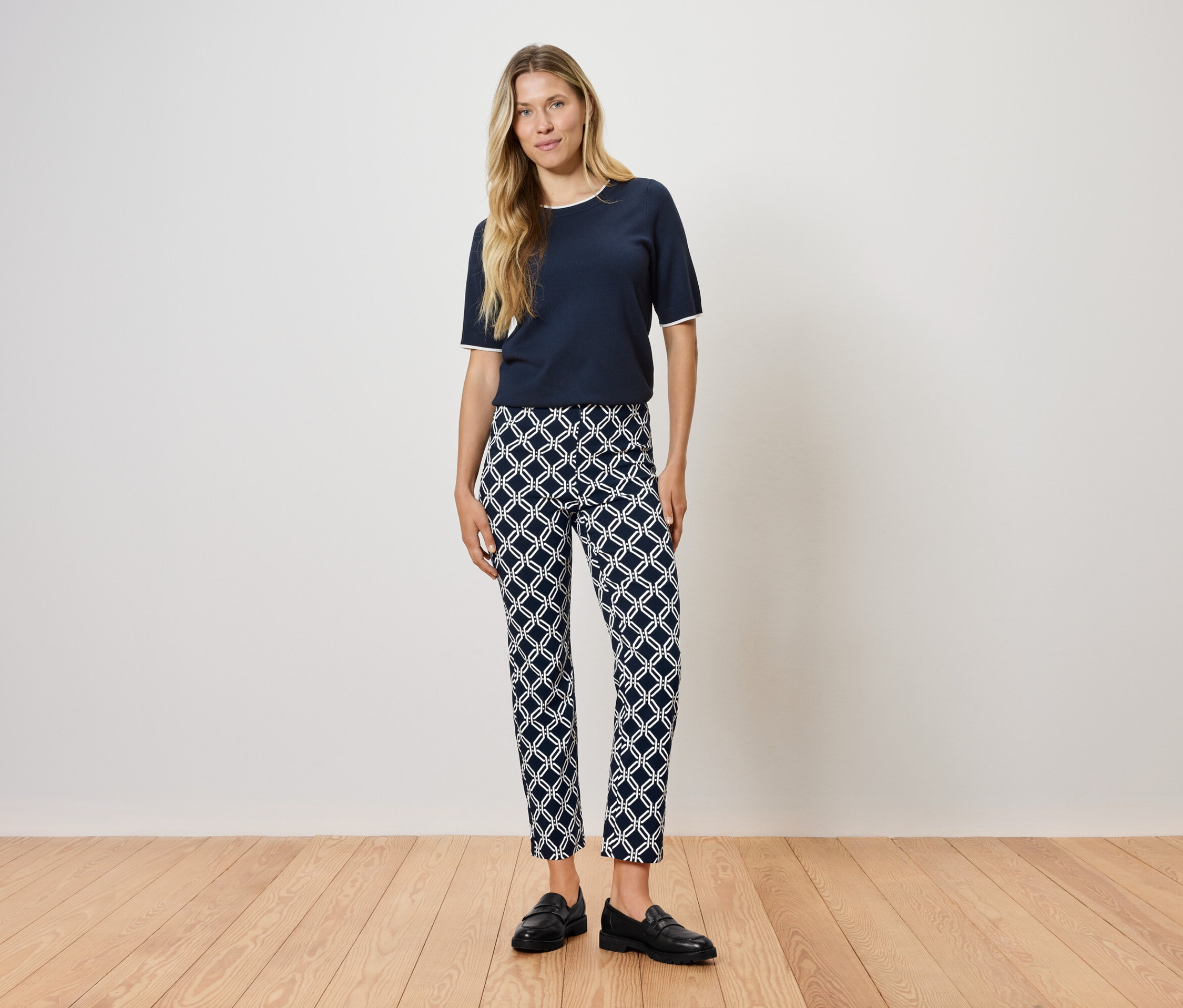 Frau steht auf Holzboden und trägt ein dunkelblaues T-Shirt und gemusterte Hosen.