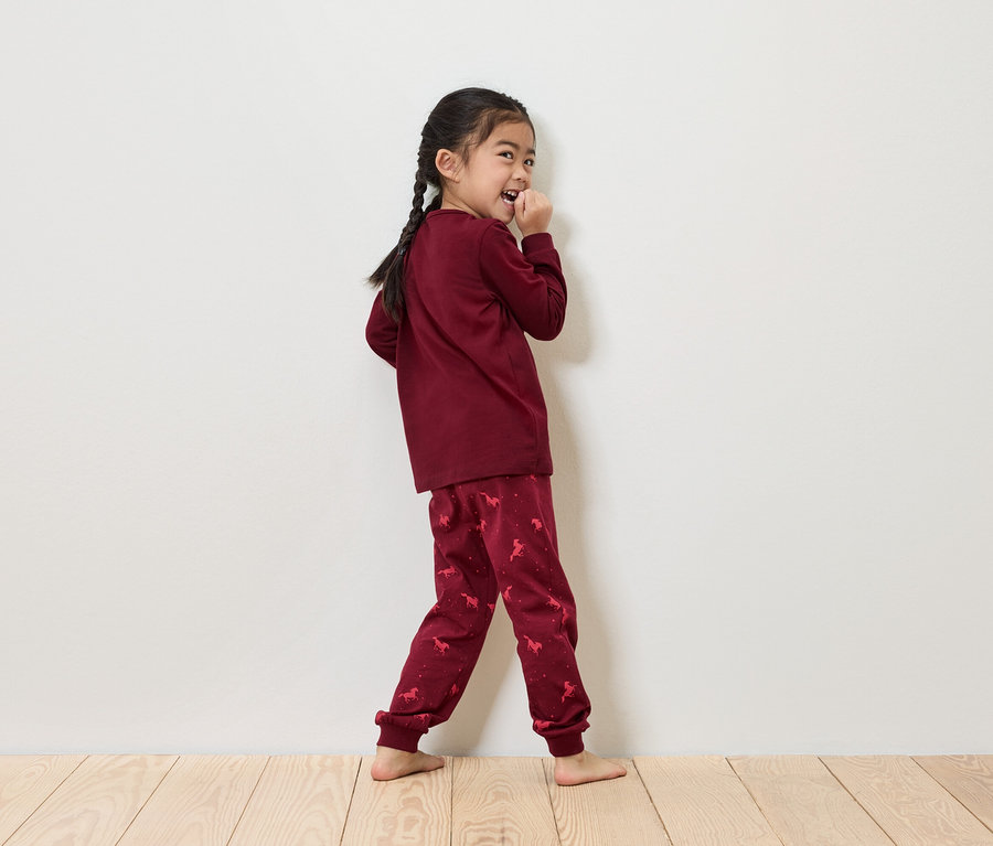 Mädchen mit Zöpfen in bordeauxfarbenem Einhorn-Pyjama steht vor einer weißen Wand.