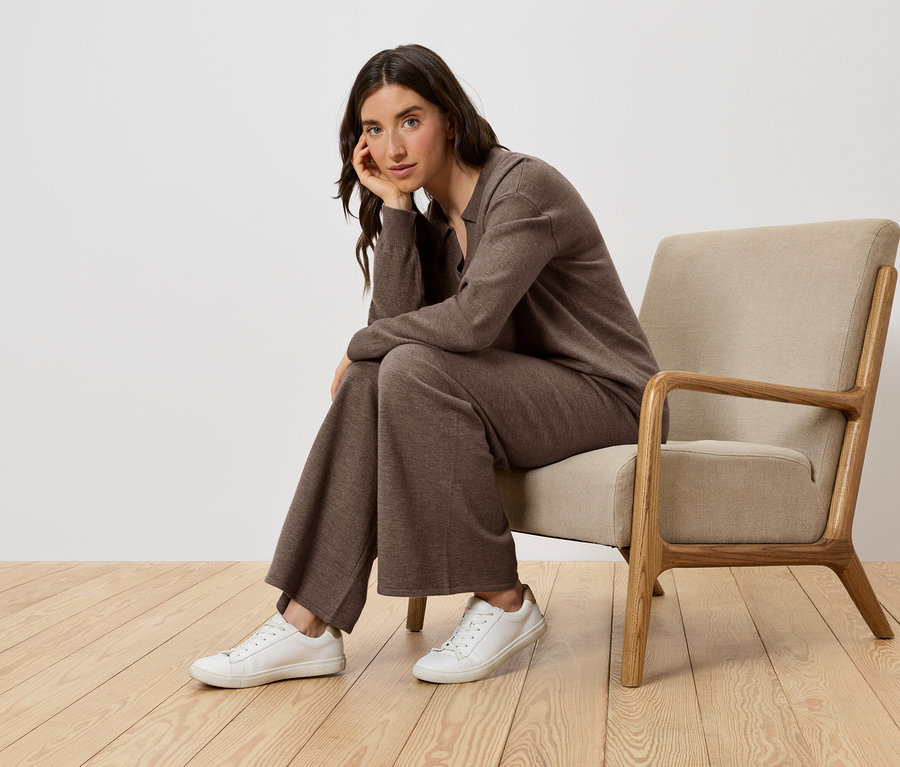 Frau sitzt in einem Sessel, trägt braune Kleidung und weiße Schuhe.