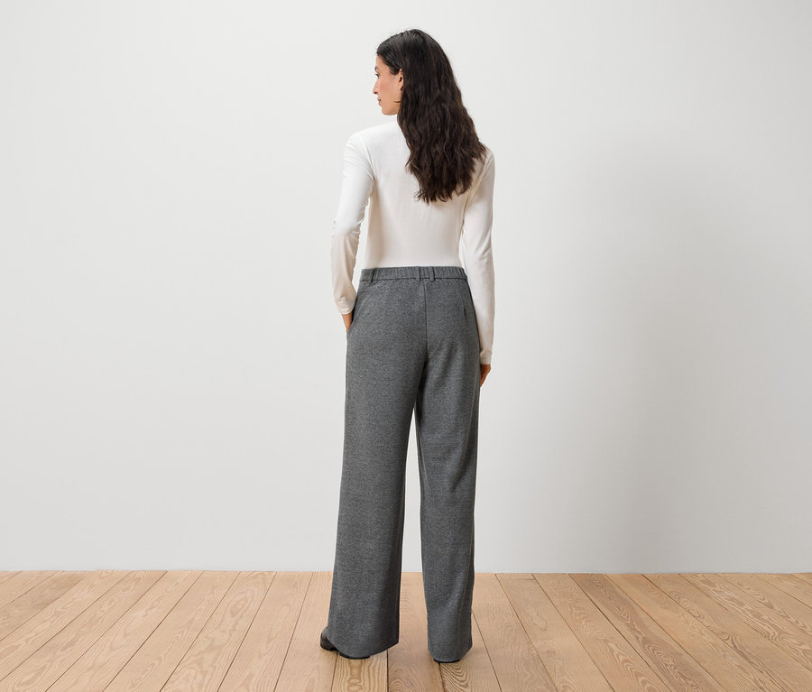 Frau mit dem Rücken zur Kamera trägt ein weißes Langarmshirt und eine graue Hose.