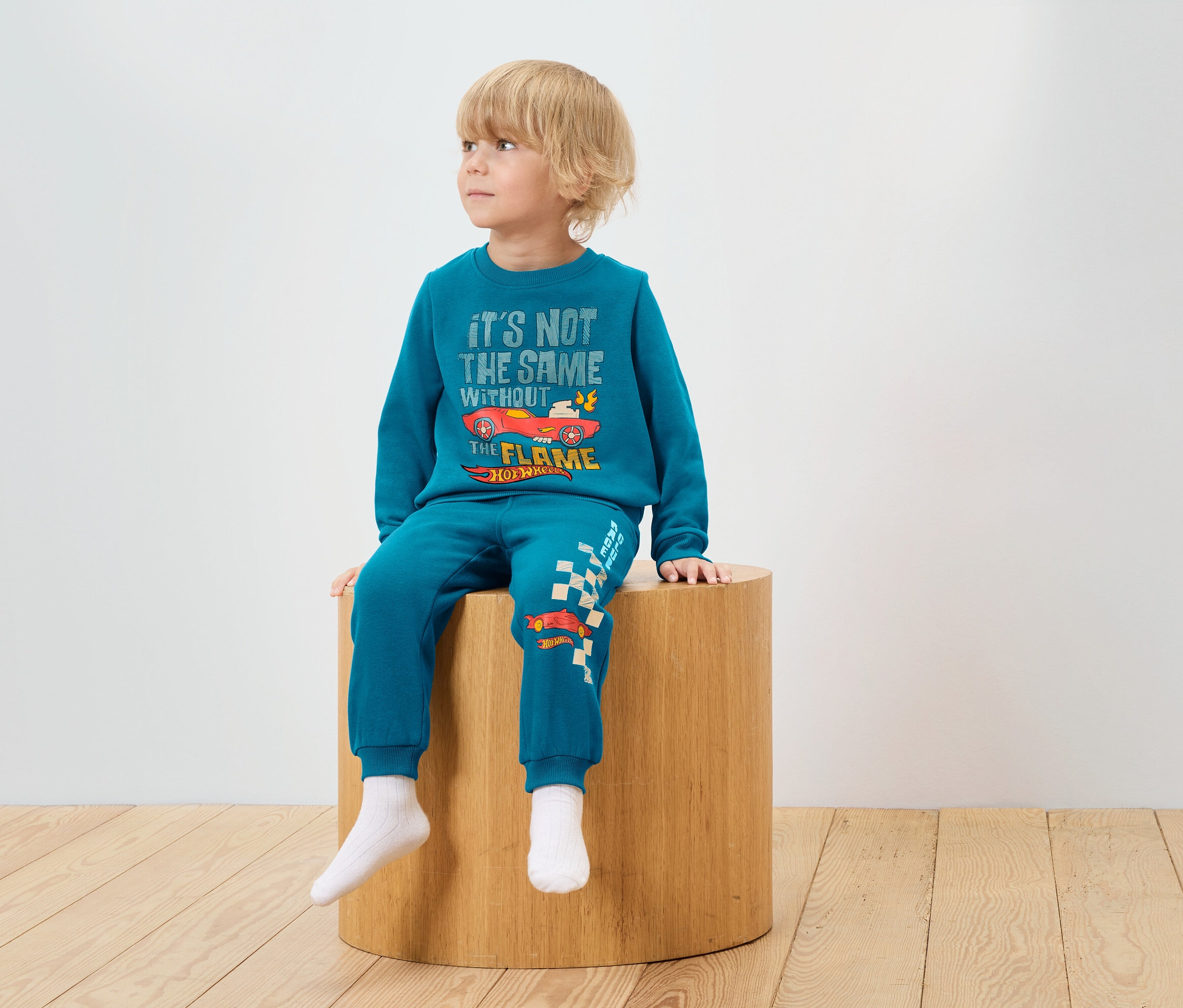 Ein Junge sitzt auf einem Holzhocker und trägt einen blauen Trainingsanzug mit Hot Wheels-Aufdruck.