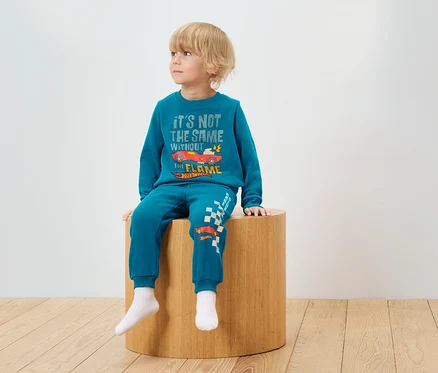 Ein Junge sitzt auf einem Holzhocker und trägt einen blauen Trainingsanzug mit Hot Wheels-Aufdruck.