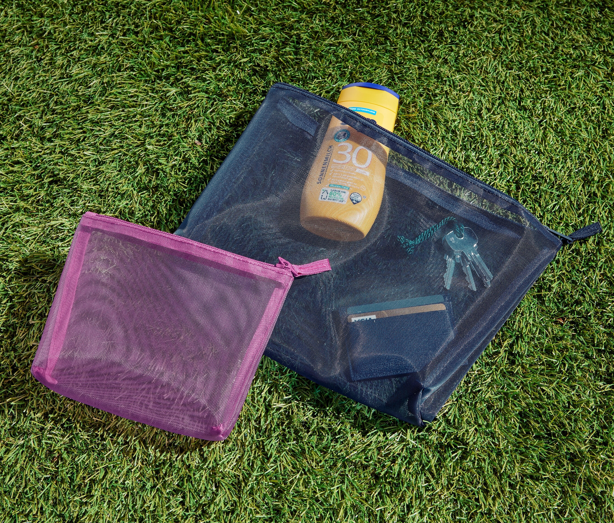 Zwei Aufbewahrungstäschchen liegen auf dem Gras. In einem davon befinden sich Sonnencreme, Schlüssel und eine Karte.