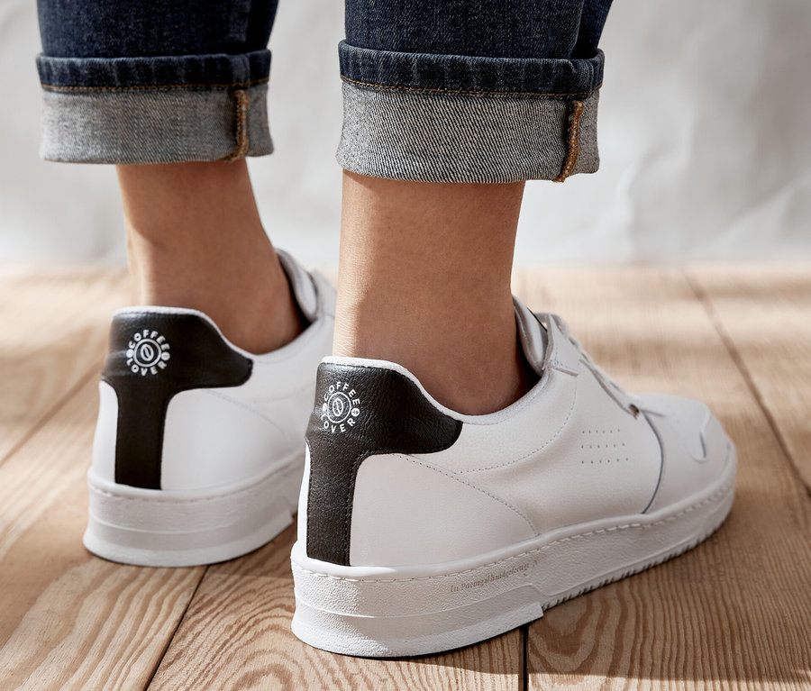 Jemand steht auf einem Holzboden und trägt weiße Tchibo x VLACE Coffee Lover Sneaker.