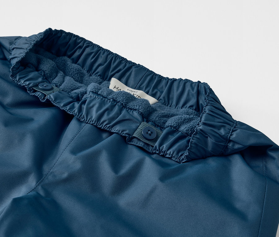 Detailaufnahme einer blauen Kinder-Regenhose mit Mitwachsfunktion und Knöpfen am Bund.