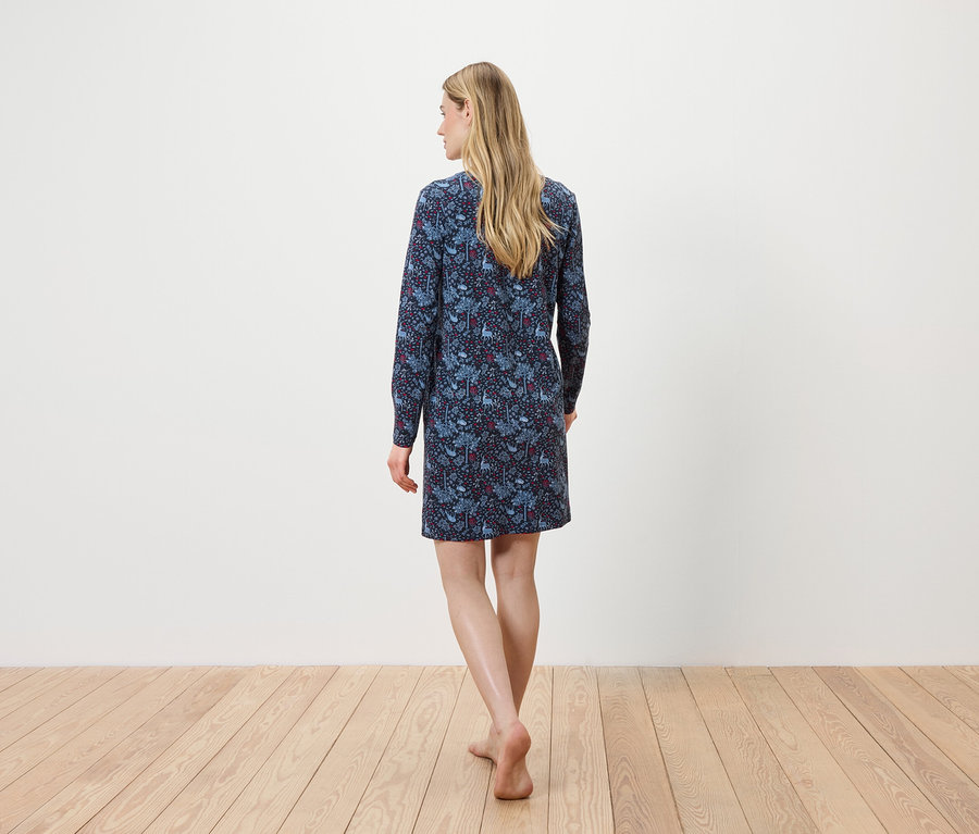 Frau mit dem Rücken zur Kamera geht barfuß auf einem Holzboden und trägt ein blaues, langärmeliges Blumenkleid.