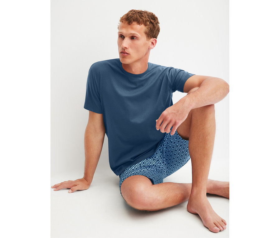 Mann sitzt auf dem Boden und trägt einen blauen CALIDA Relax Streamline 2 Shorty-Pyjama mit gemusterten Shorts.