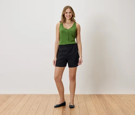 Frau mit grünem Top und schwarzen Shorts steht auf einem Holzboden vor einer weißen Wand.